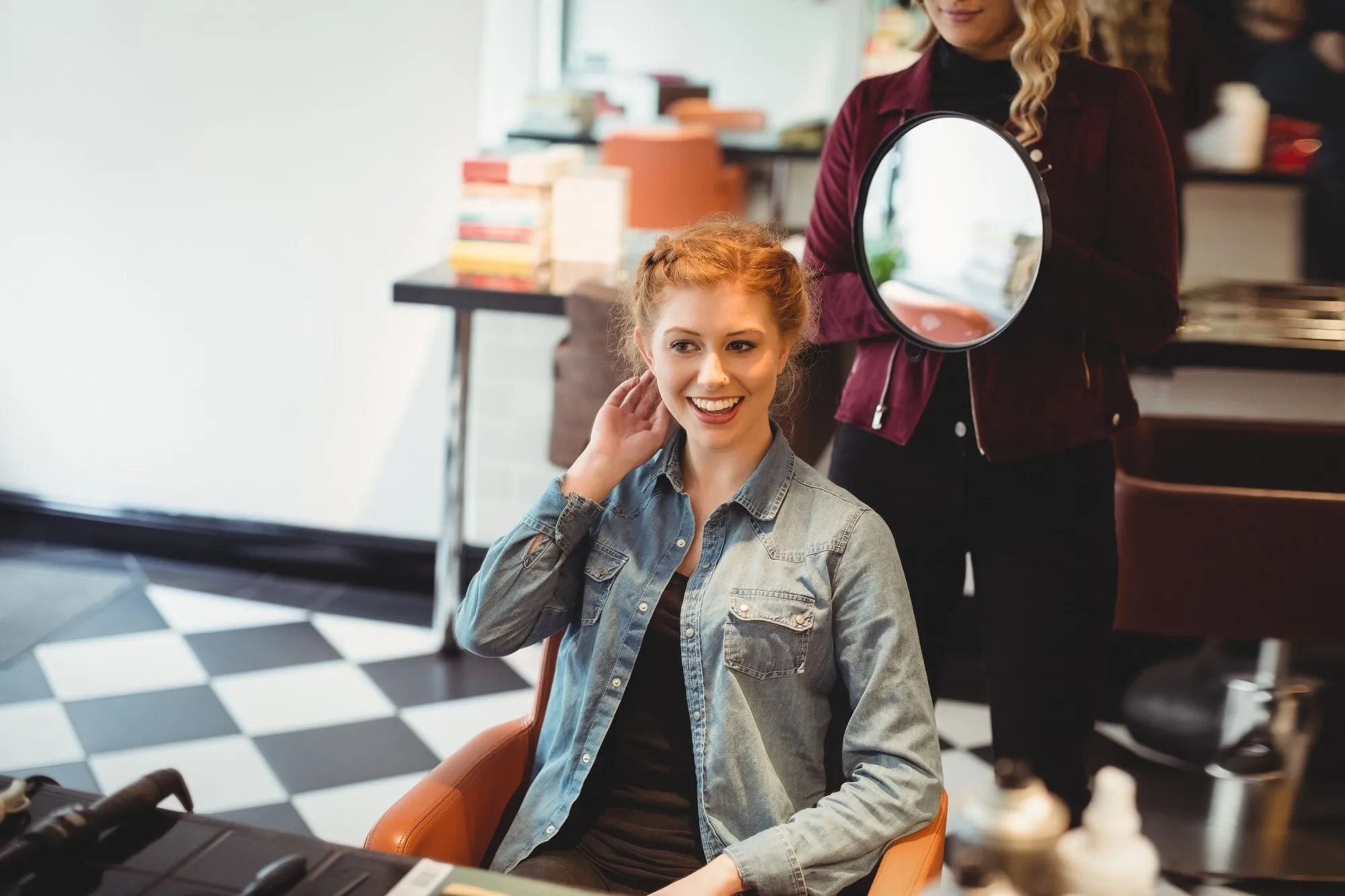 Glimlachende vrouw met opgestoken rood haar zit in een kappersstoel, terwijl de specialist achter haar een ronde handspiegel omhoog houdt.